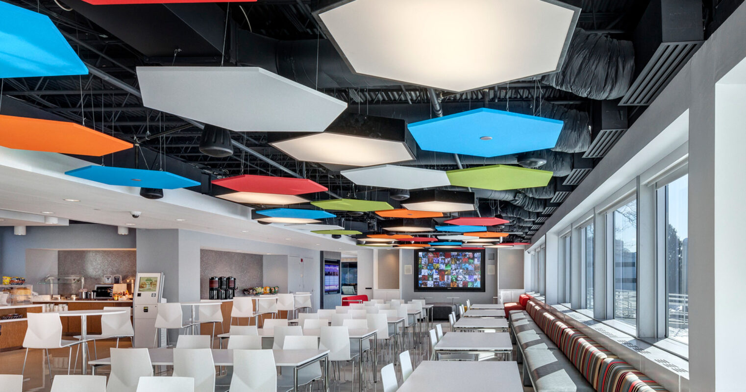 Vibrantly colored geometric acoustic panels are suspended from an exposed ceiling over a modern cafeteria space, complemented by a variety of seating options and natural light from floor-to-ceiling windows.