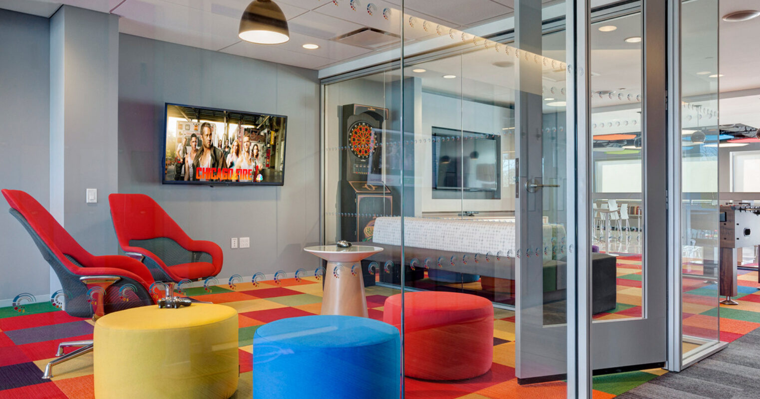 Modern office lounge with vibrant multicolored carpet, red ergonomic chair, and yellow and blue poufs. Glass walls provide transparency, with playful dartboard accentuating the creative atmosphere.