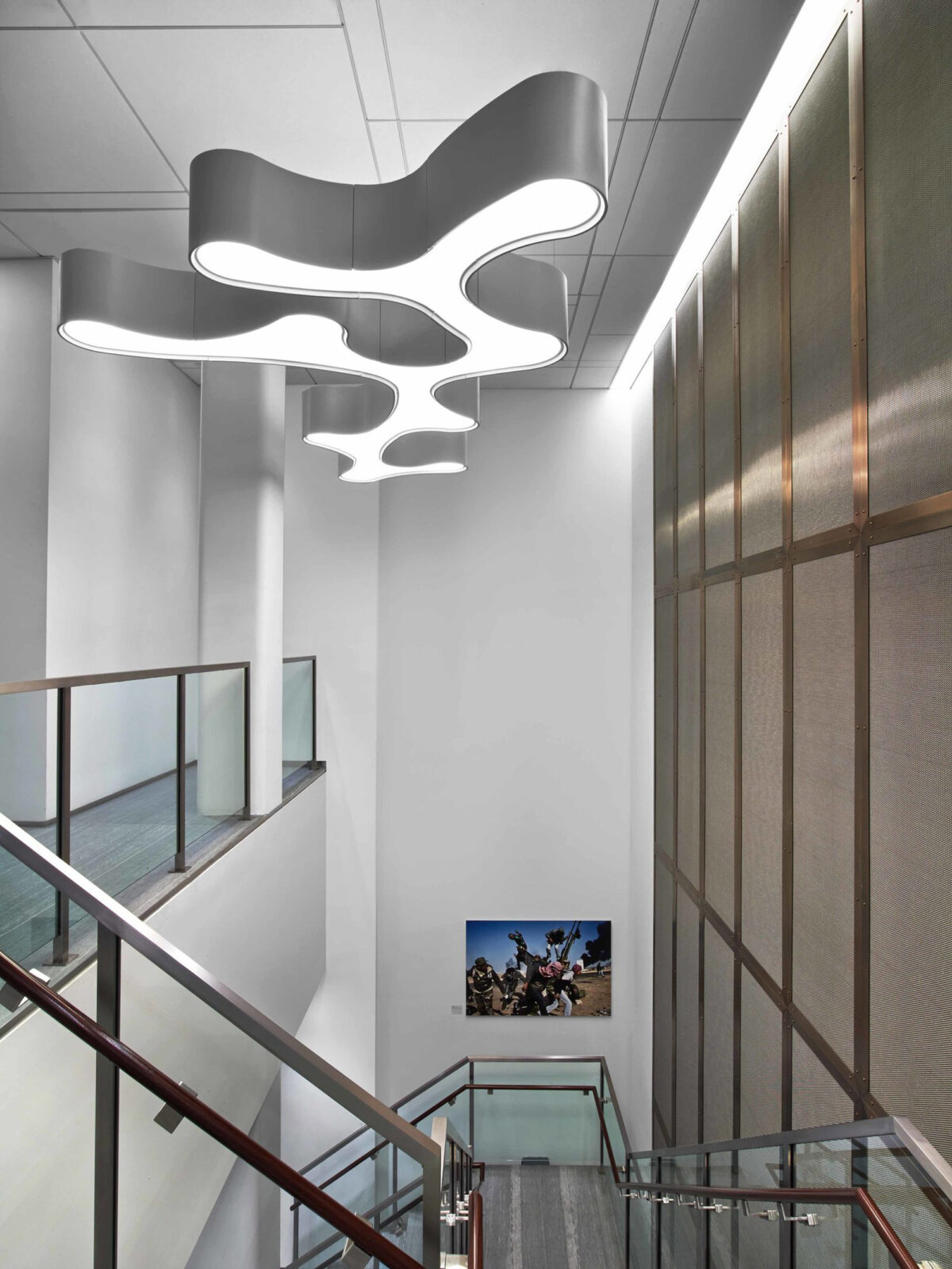 A modern interior with flowing, sculptural white LED light fixture suspended above a staircase with stainless steel handrails, glass balustrades, and wood-paneled walls, accented with a large photograph of motorcyclists on the lower landing wall.