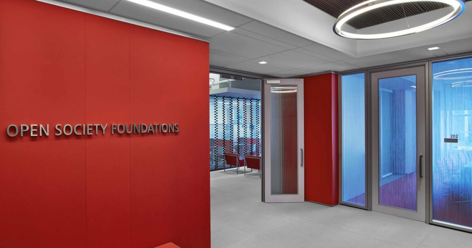 Modern office corridor featuring a vibrant red wall with the text 'OPEN SOCIETY FOUNDATIONS', complemented by a sleek red bench. The space is defined by contemporary lighting fixtures and two-toned flooring, leading to a glass door marked '202'.