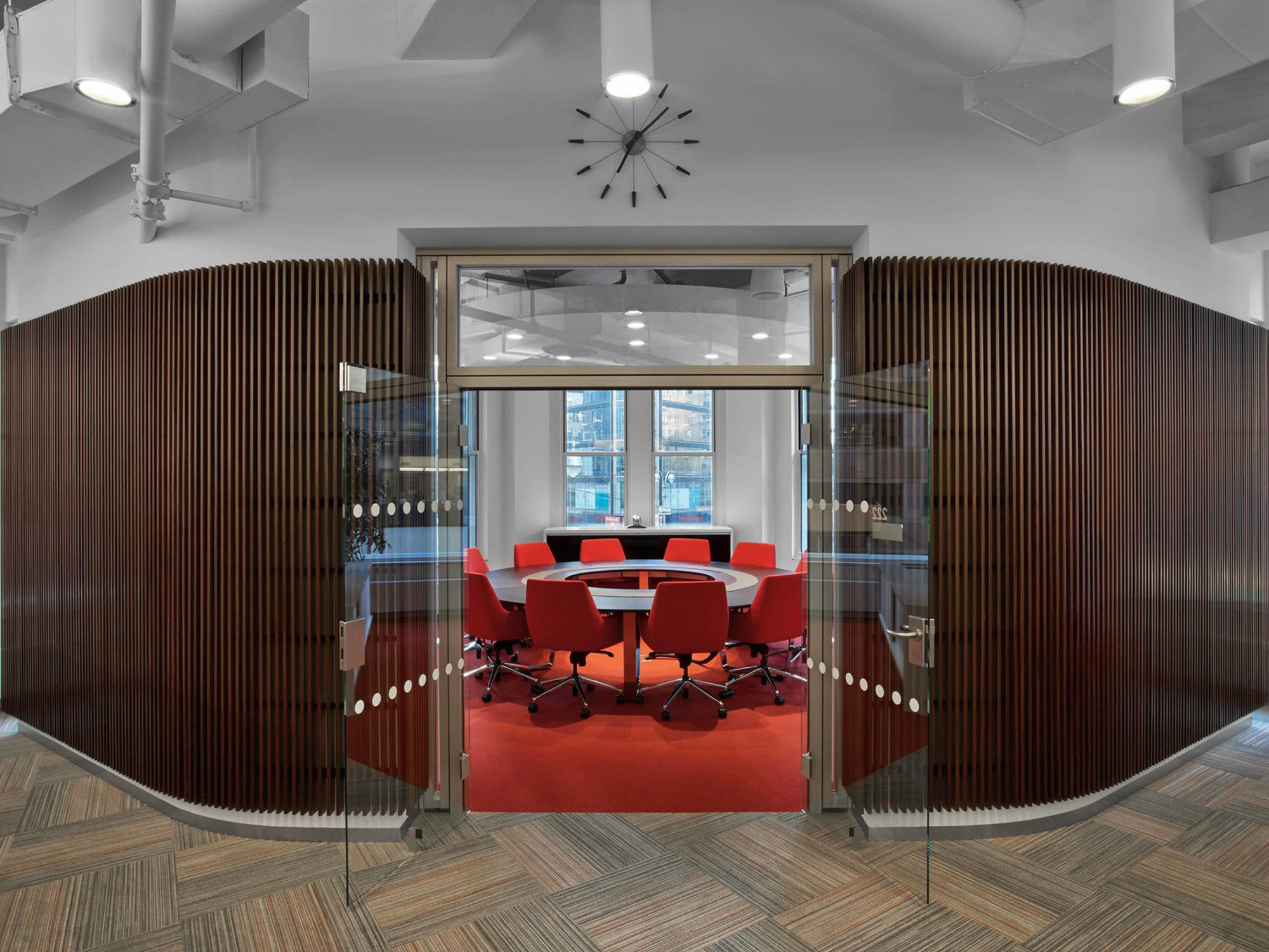 Modern office conference room with curved wooden slat partitions, red chairs around a circular table, and a herringbone-pattern floor complementing the neutral color palette. Clear glass doors offer a glimpse into the collaborative space, while a starburst ceiling light fixture adds a mid-century touch.