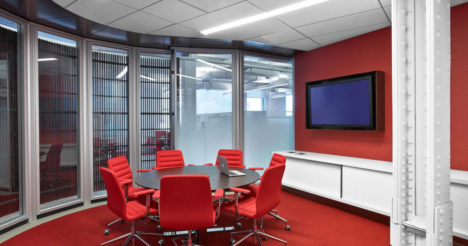 Modern conference room featuring a sleek, round table with vibrant red chairs, set against a backdrop of red carpet, stark white walls, and expansive windows with adjustable horizontal blinds. A flat-screen monitor adorns one wall, complementing the room's contemporary aesthetic.