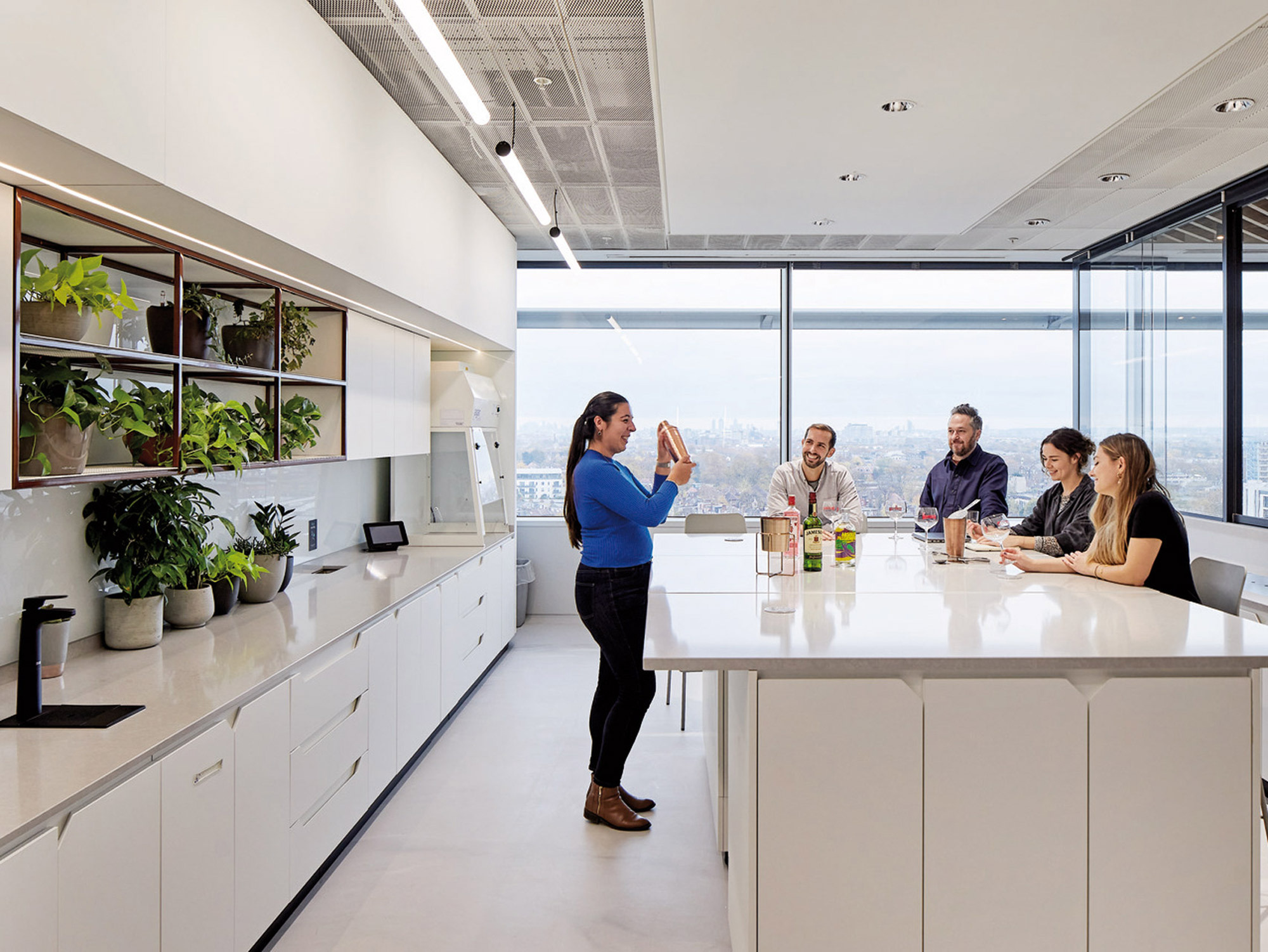 Modern office kitchen and break area with minimalist white cabinetry, expansive windows offering city views, and abundant natural light. Green plants add a vibrant touch to the clean, functional design. A group of professionals engage in a casual meeting around the central island.