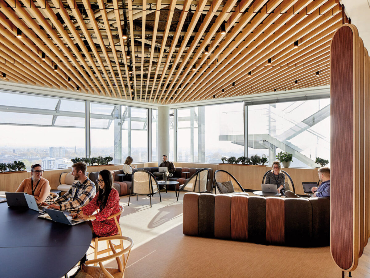 Vaulted ceiling with linear wooden slats shades a modern office lounge. Varied seating arrangements with woven accents promote collaboration and individual work amidst floor-to-ceiling windows showcasing a cityscape. Natural light complements the warm, earthy tones of the furniture and fixtures.