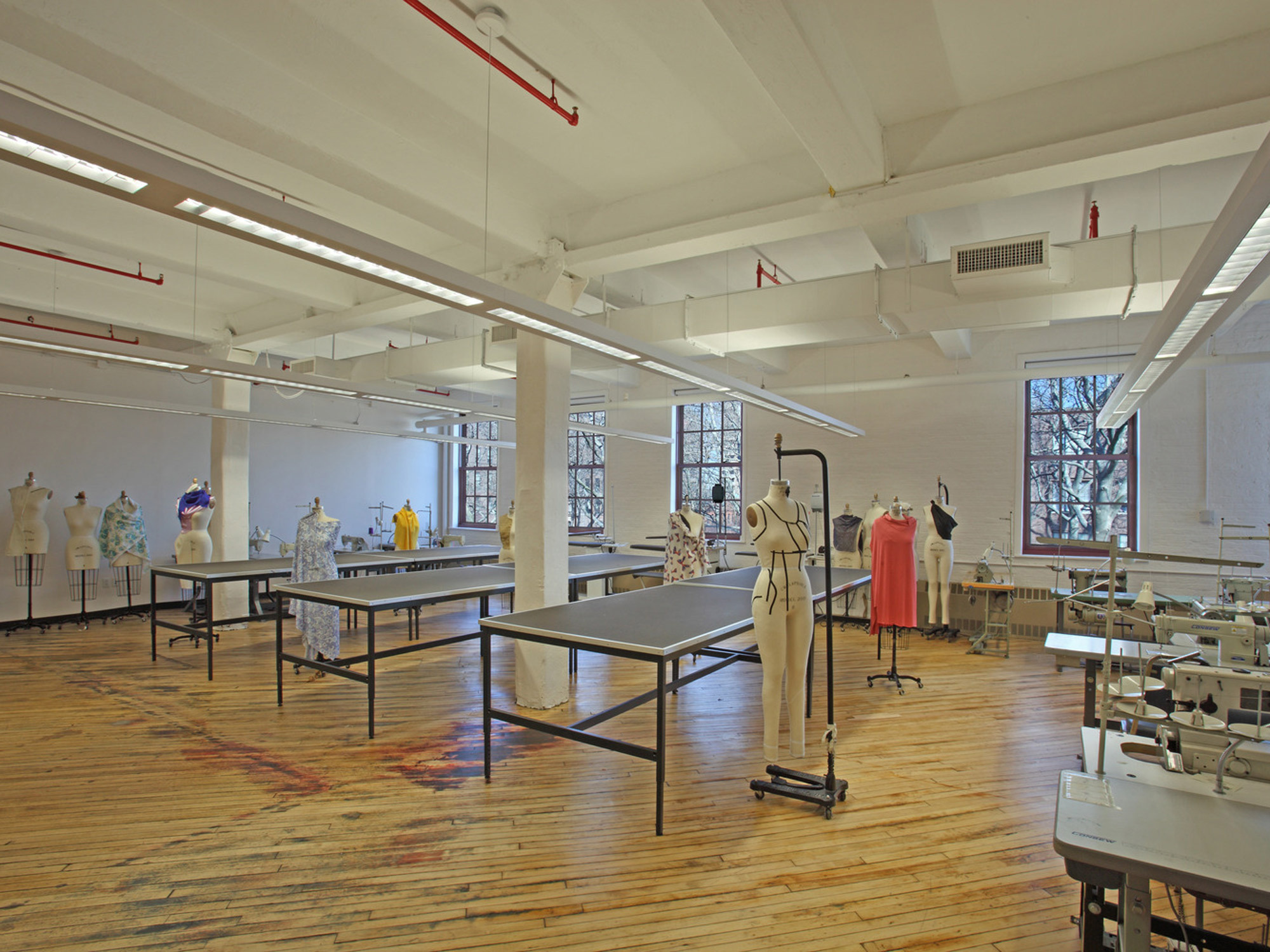 Spacious fashion design studio with bright natural light, featuring stark white walls, high ceilings, and exposed beams. Workstations with sewing machines and dress forms are organized for functionality, while splashes of color on the distressed wooden floor add character to the creative workspace.