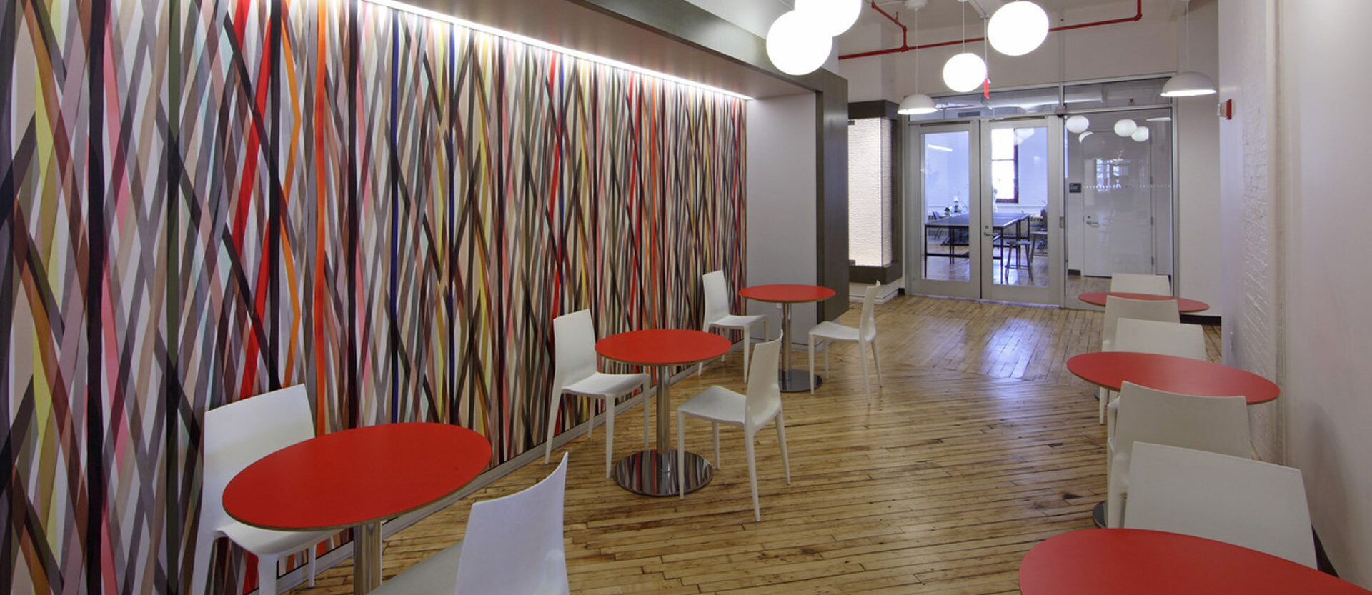 Modern office break room featuring a vibrant, geometric patterned accent wall, white contemporary chairs with red cushions, round white pendant lights, and natural wood flooring adding warmth to the minimalist design. The space is bright, inviting, and encourages informal collaboration.