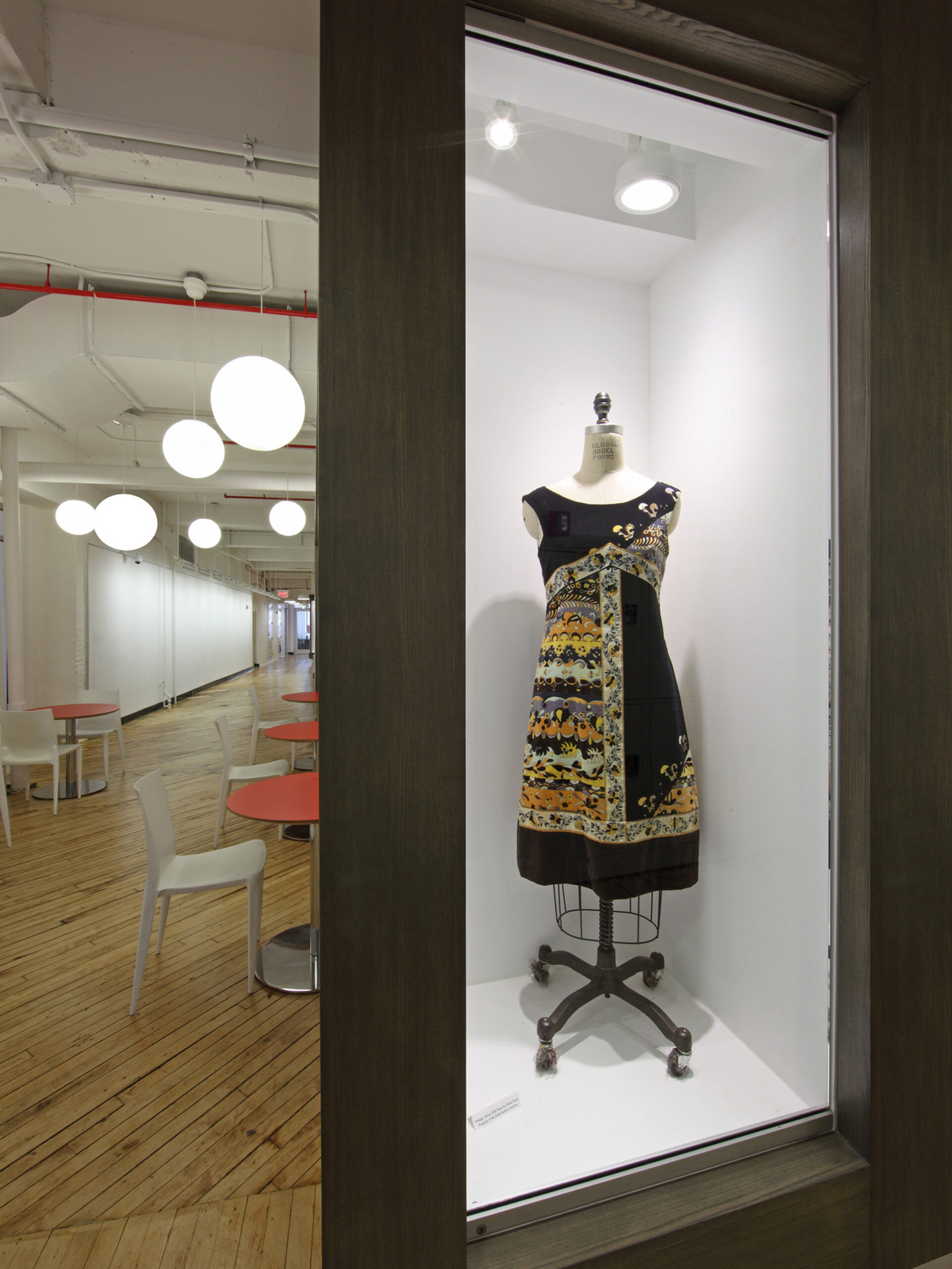 Vintage mannequin displaying an ornate dress, framed by a minimalist storefront window, with the backdrop of a modern, open-concept interior featuring sleek furniture and bold red ceiling-mounted pipes.