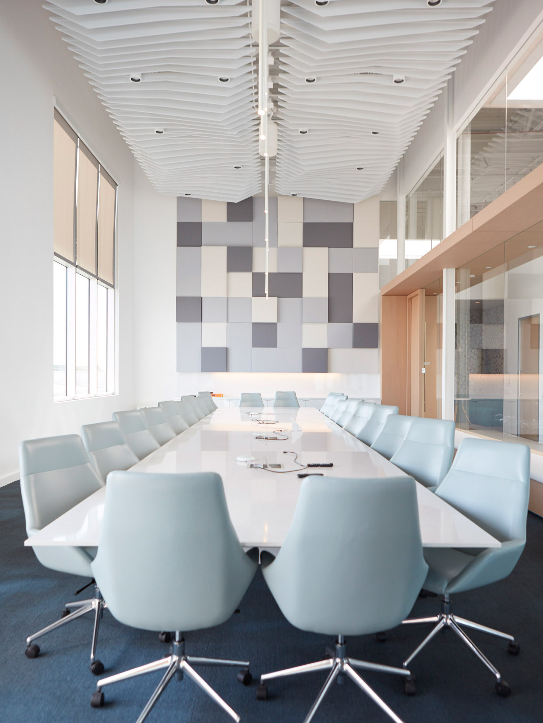 Modern conference room featuring a long white table with rounded edges surrounded by ergonomic aqua chairs. The space is defined by a geometric gray and white wall mosaic and a sculptural ceiling with dynamic wave patterns enhancing the room's acoustic qualities. Natural light filters through ample windows.