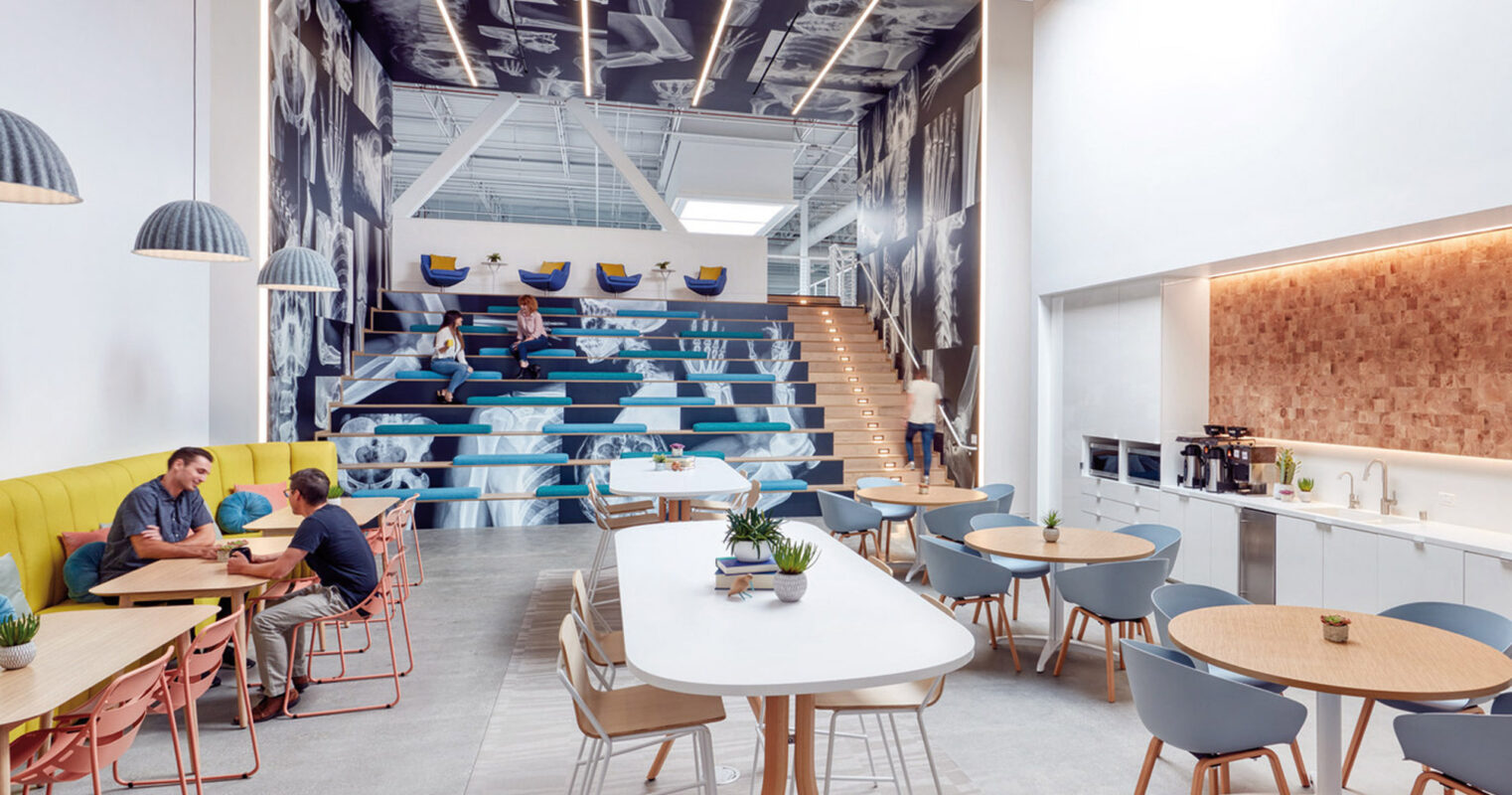Modern office break room featuring eclectic furnishings, pastel hues, and a staircase doubling as auditorium seating with a large monochrome mural enhancing the vertical space. Natural light brightens wood tonality and reflective surfaces, fostering a dynamic communal ambiance.
