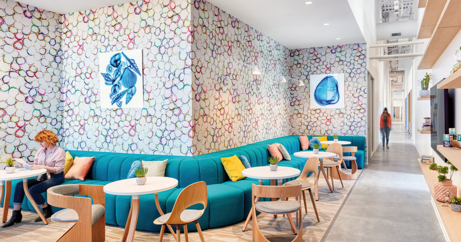 Bright, modern interior featuring a curved turquoise sofa against a vibrant, organically patterned wall. Natural wood accents and simple white tables create a welcoming atmosphere, complemented by whimsical jellyfish art prints. The space is well-lit, emphasizing the playful yet sophisticated design aesthetic.