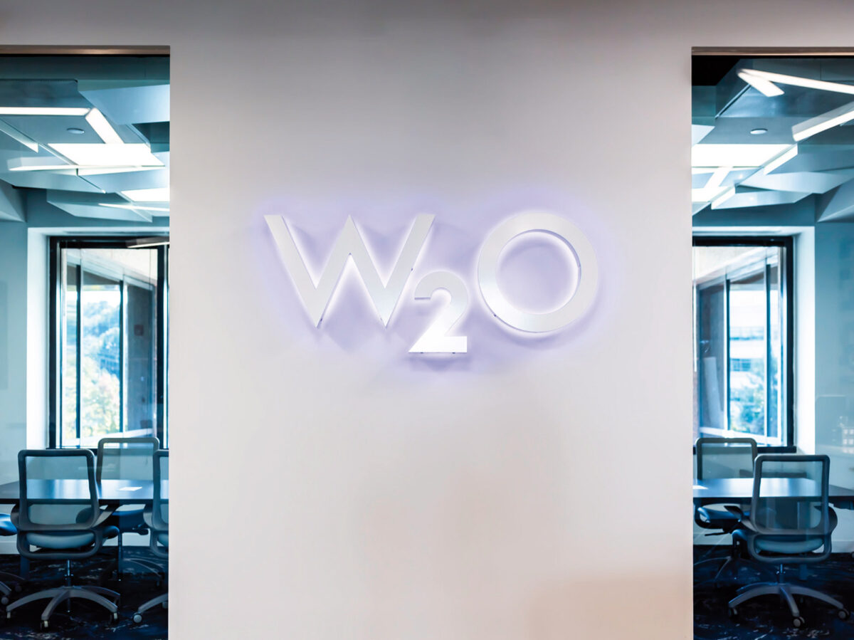 A minimalist office interior with a prominent backlit logo affixed to a white wall, flanked by transparent glass paneled conference rooms with ergonomic furnishings.