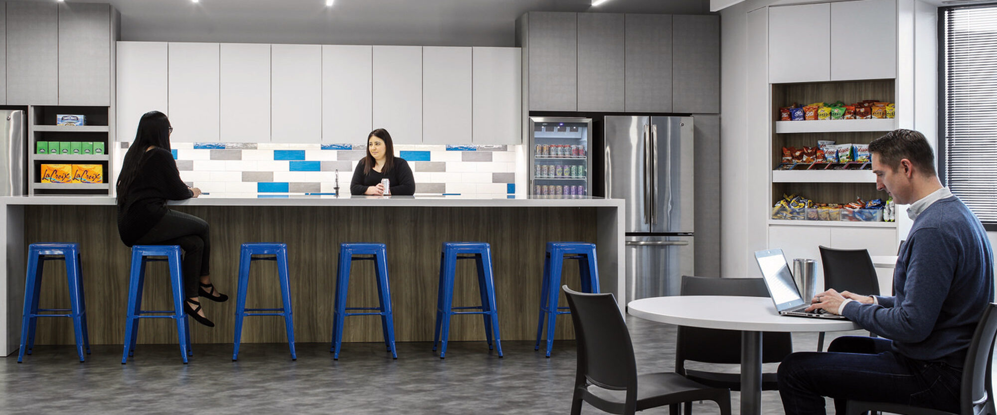 Modern office break room features a monochrome color palette with vibrant blue bar stools, providing a pop of color. Integrated lighting accentuates the textured tile backsplash. Employees engage around the functional kitchenette and workspace, fostering a collaborative environment.
