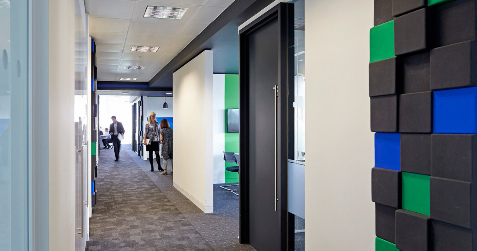 Modern office interior with a vibrant blend of white walls and black, blue, green acoustic panels. Sleek glass doors separate workspaces, while gray carpeted floors offer a neutral path. Contemporary lighting enhances the functional yet aesthetically pleasing business ambiance.