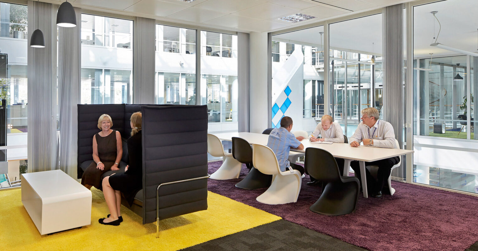 Modern office breakout area featuring high-back acoustic seating for privacy, vibrant yellow and purple floor mats for zoning, and white minimalistic side tables. The glass walls provide transparency, while pendant lighting adds to the contemporary ambiance.