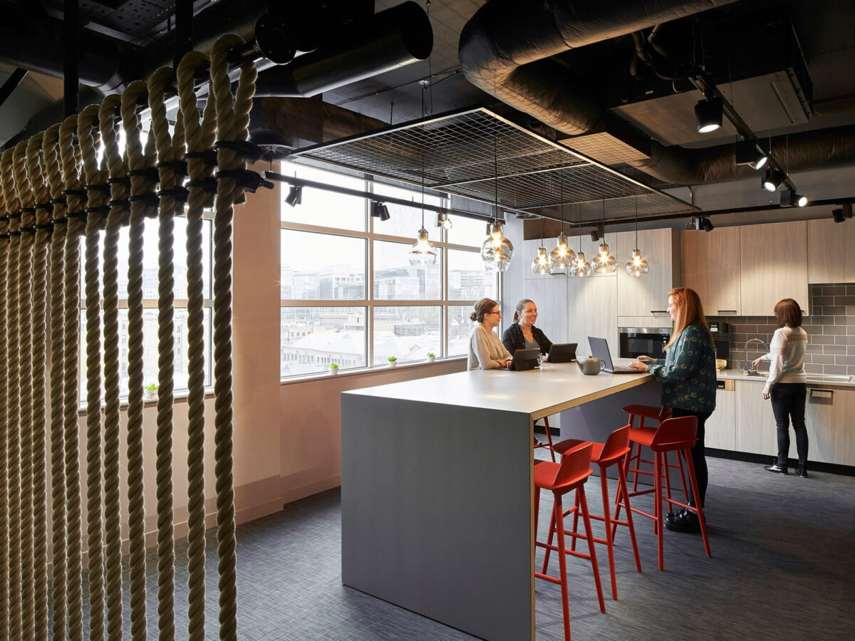 Modern open-plan office space featuring an industrial chic aesthetic with exposed ductwork, pendant lights above a high-top communal table with red stools, woven room dividers, and employees engaged in conversation.