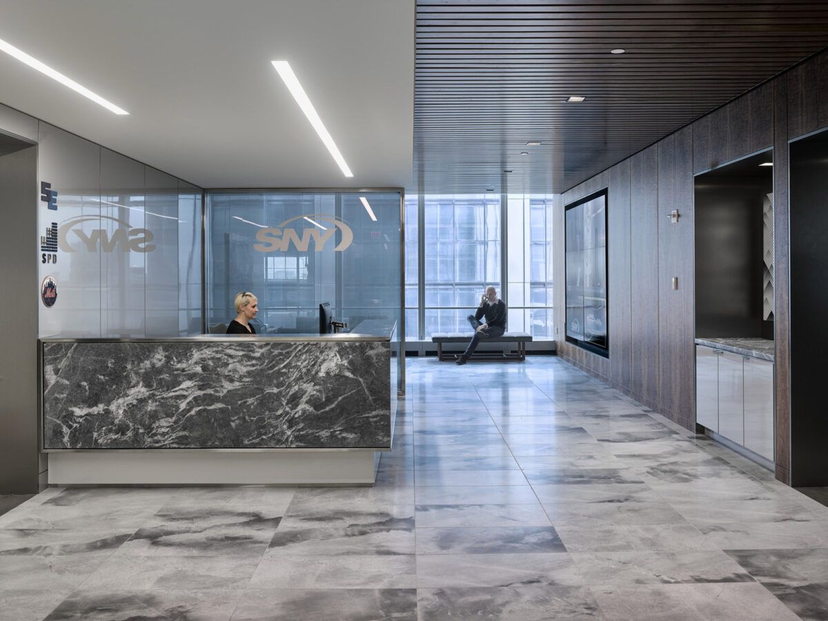 Modern office lobby featuring a sleek reception desk with marble patterns, flanked by translucent glass walls and a wooden slat accent wall. The flooring is light-toned, polished stone, complementing the neutral, sophisticated color scheme of the space.