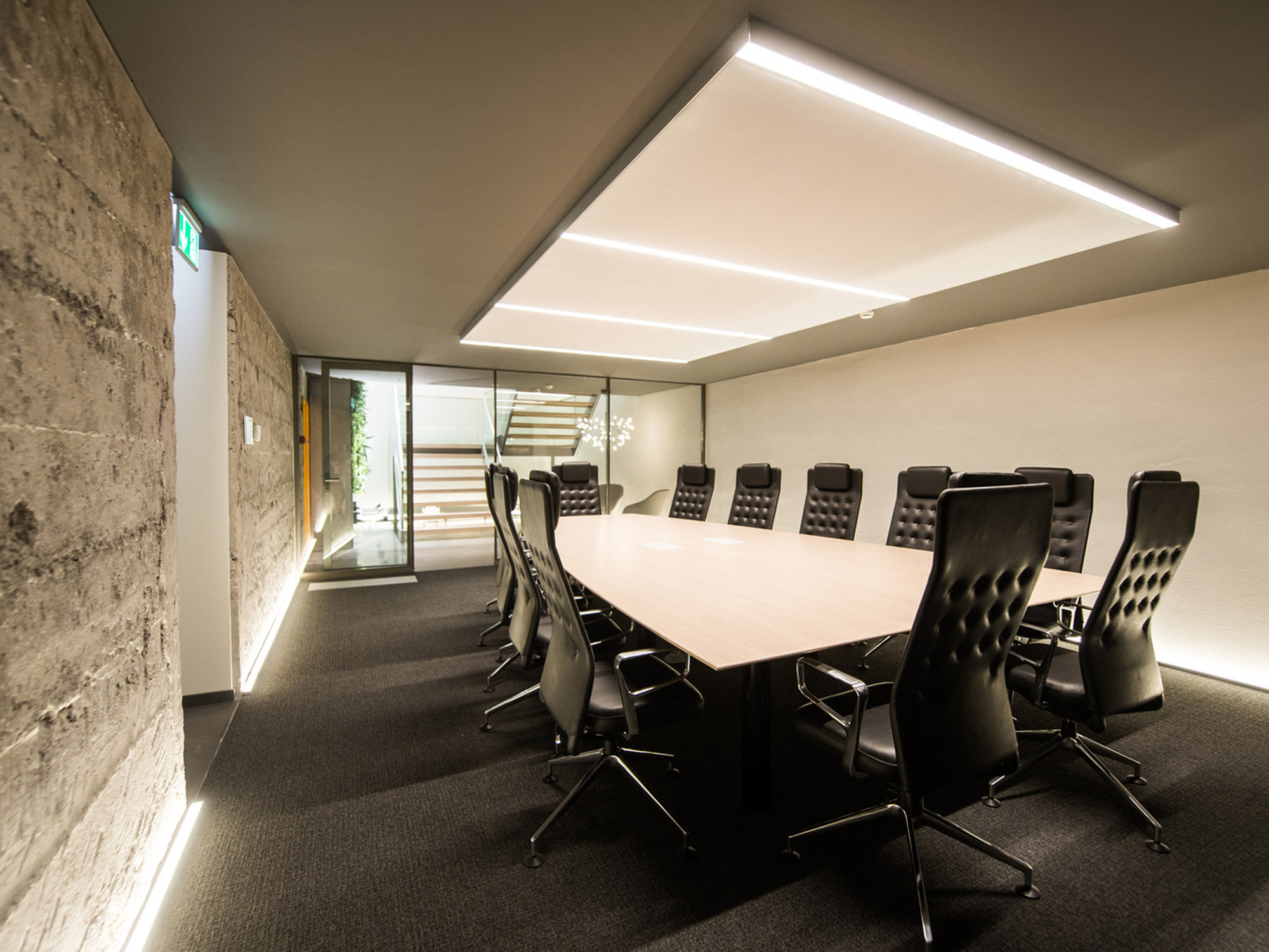 Spacious modern conference room featuring an elongated, polished wooden table, surrounded by black leather chairs, accentuated by a striking linear suspended LED lighting fixture and textured concrete walls.