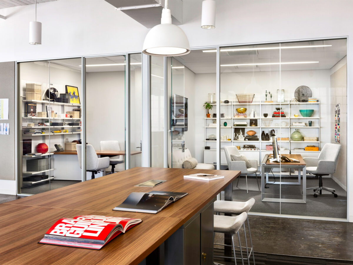Modern office interior with clean lines featuring a polished wooden conference table, white ergonomic chairs, and glass partition walls. The well-lit space showcases shelving with decorative items, reflecting a blend of functionality and aesthetic appeal.