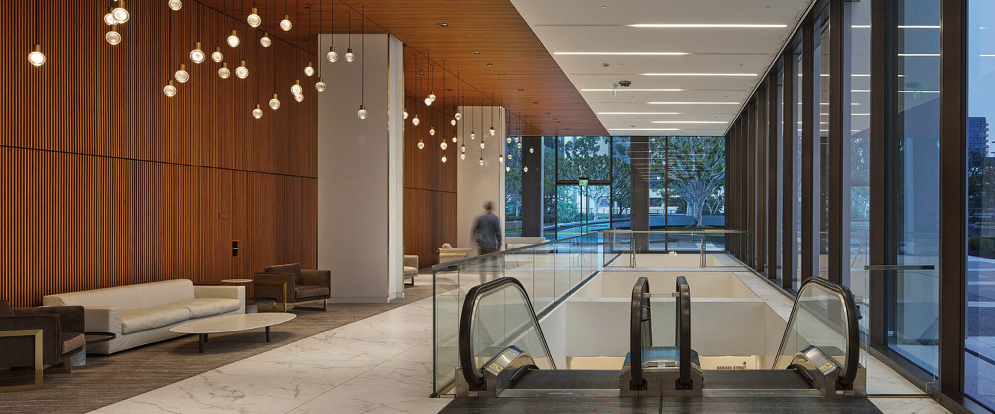 Modern commercial lobby featuring warm wood panel ceiling, recessed lighting, and clusters of unique filament pendant lights. White benches complement the marble flooring, while expansive windows afford a view of the urban setting outside.