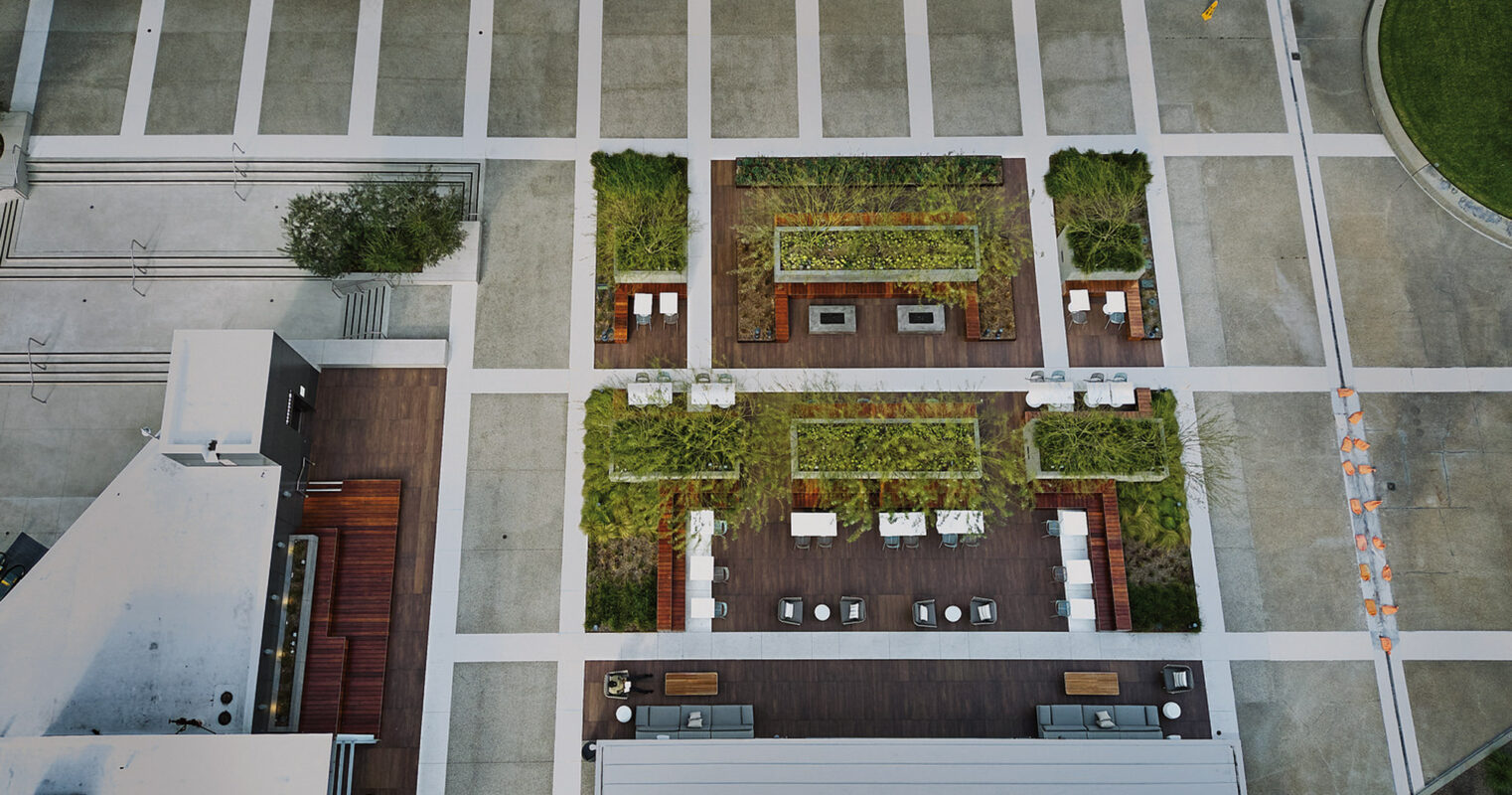Bird's-eye view of a rooftop garden with arranged green spaces among wooden decking, flanked by contemporary outdoor furniture and surrounded by urban architectural elements. Design seamlessly integrates natural and built environments, providing a relaxing communal space atop a city structure.