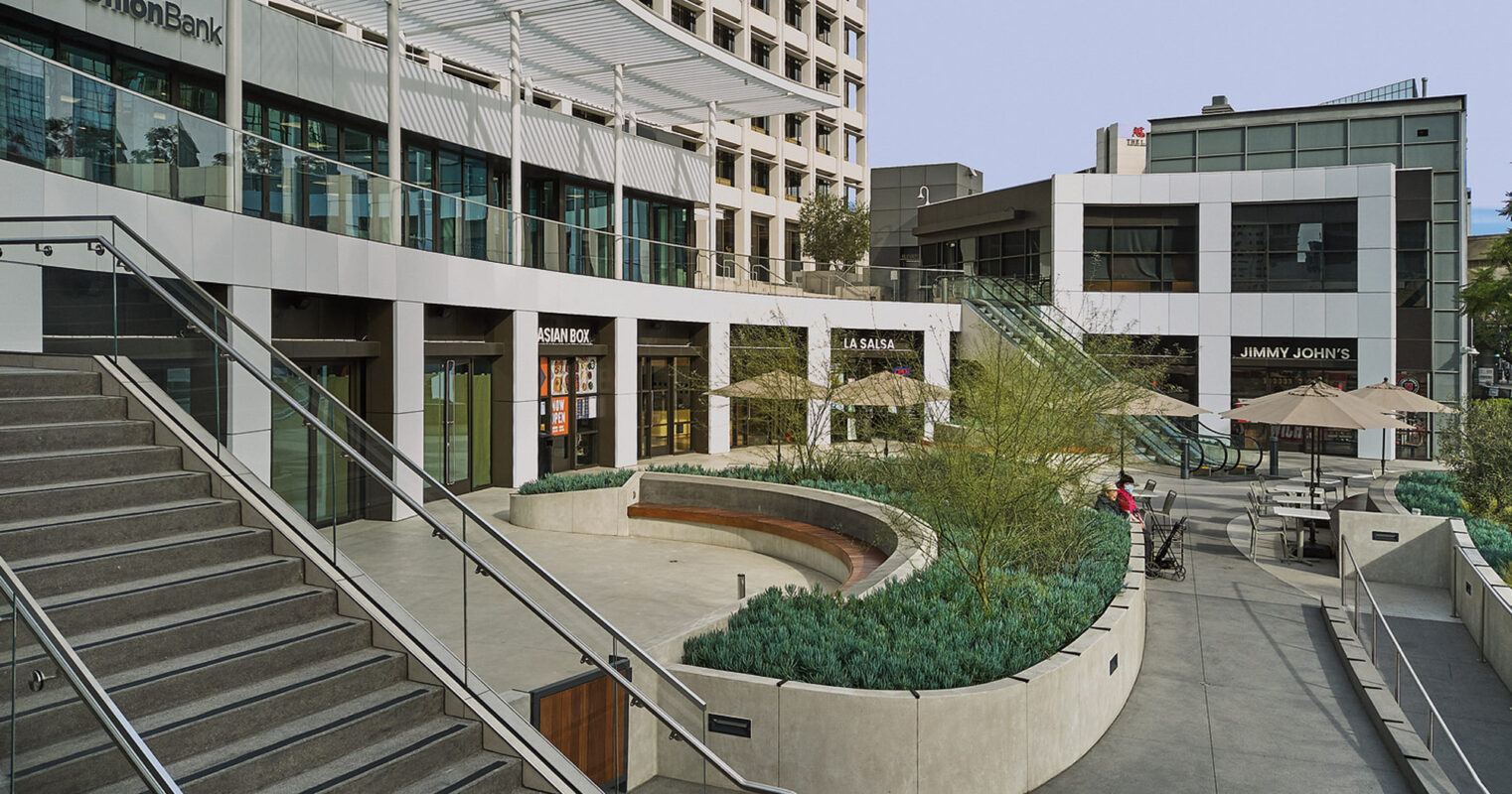 Modern urban plaza featuring tiered concrete steps leading to a central seating area with curved planters, surrounded by contemporary buildings and retail facades.