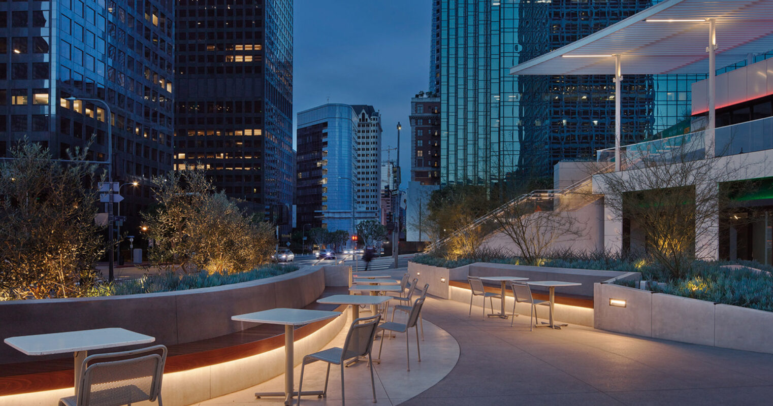 Modern urban rooftop terrace with sleek, curved benches and minimalist white tables. Soft lighting accentuates the understated elegance amidst the high-rise backdrop at twilight.