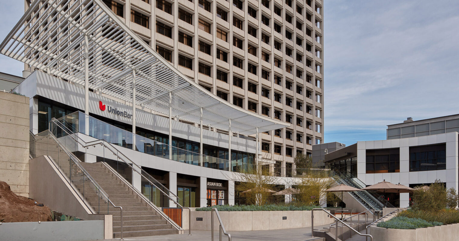Modern urban staircase leading up to an elevated plaza with geometric metal canopy, flanked by a multi-story building and landscaped areas, under a partly cloudy sky.