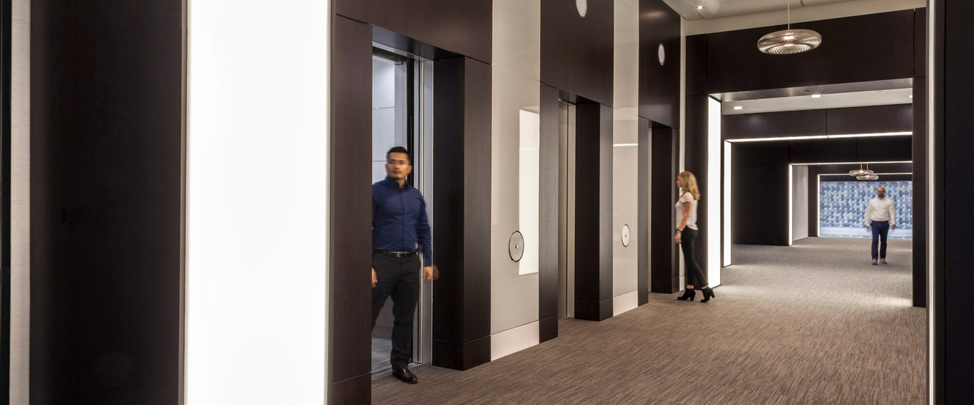 Elegant modern hallway featuring high-contrast design with dark wood panels and recessed white lighting. Geometric carpeting adds depth, while a traditional chandelier provides a classic touch against the clean lines. People casually walk, suggesting a dynamic corporate or hospitality space.