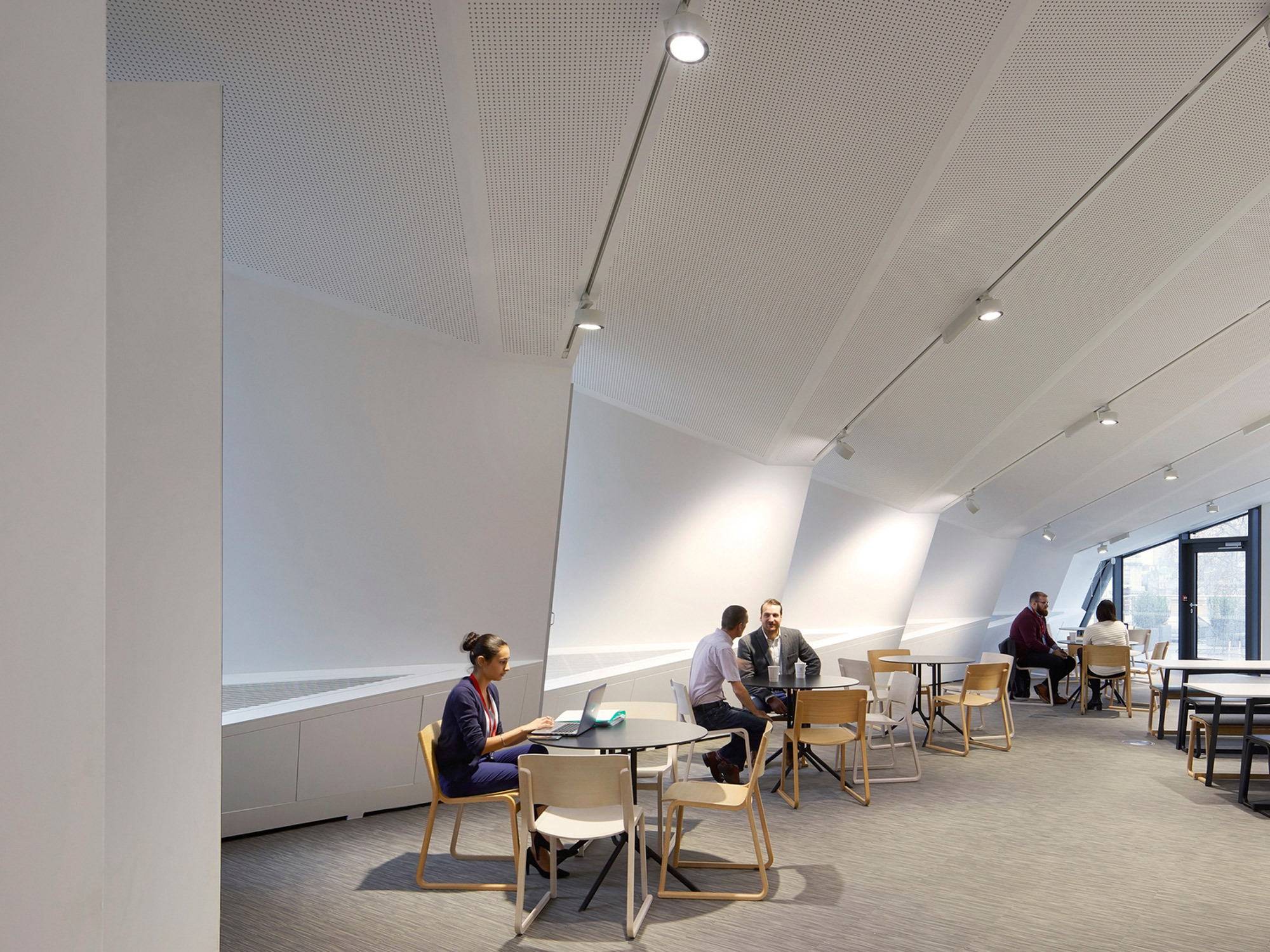 Modern office space with slanted ceilings and abundant natural light. Minimalist design features clean lines, white surfaces, and strategic lighting. Individual workstations and communal tables facilitate both focus and collaboration.