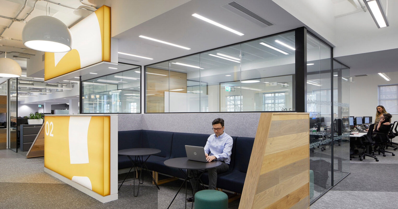 Modern workplace featuring an open floor plan with clear glass partitions, integrated yellow and blue acoustic panels, ambient pendant lighting, and ergonomic furniture. A professional works focused at a mobile laptop station against a minimalist yet vibrant design backdrop.