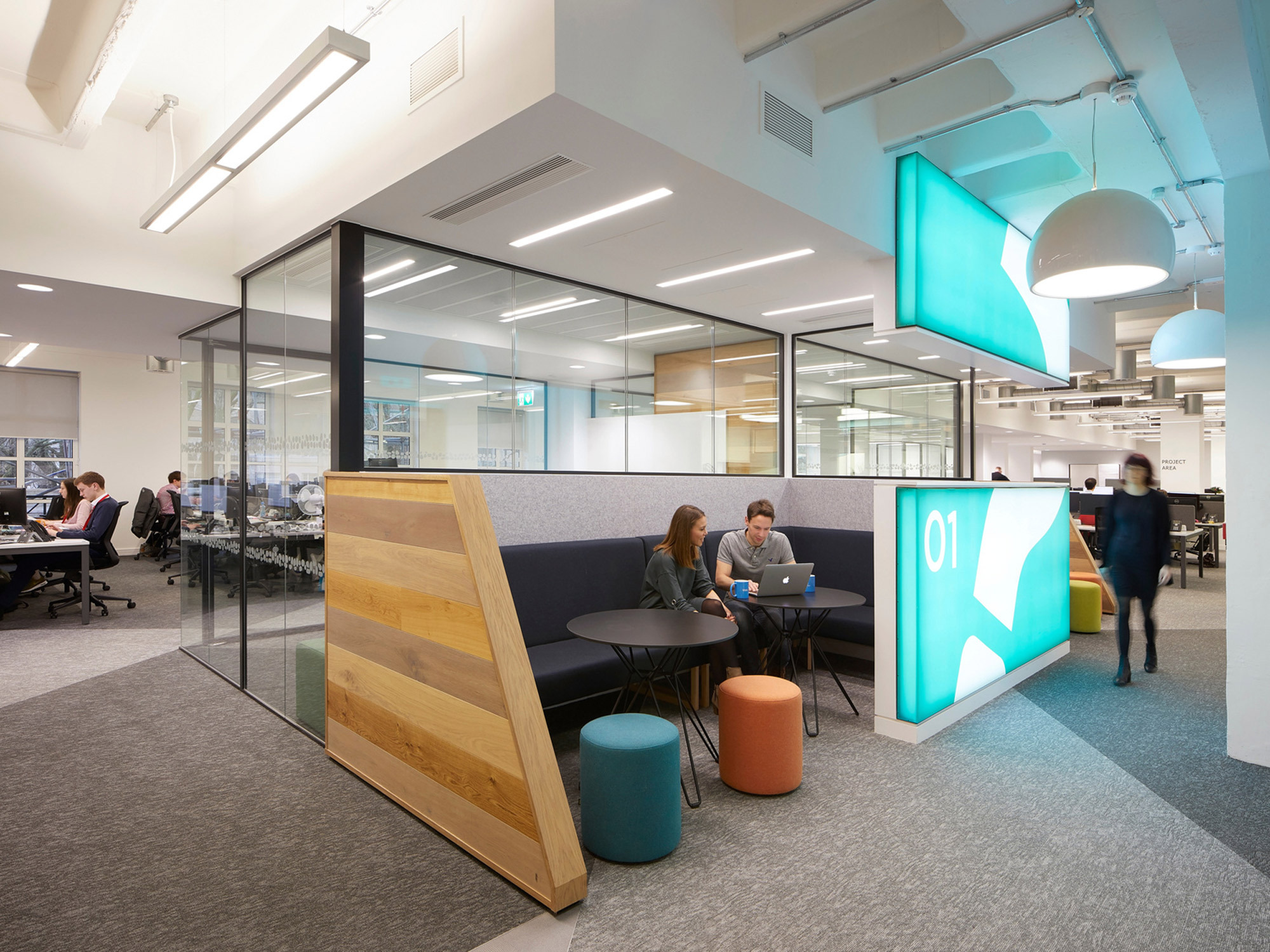 Modern office space featuring glass partition walls, an informal meeting area with colorful poufs, and dynamic lighting. A bold, illuminated numeral adds a creative touch, while employees work at clustered desks in the background.
