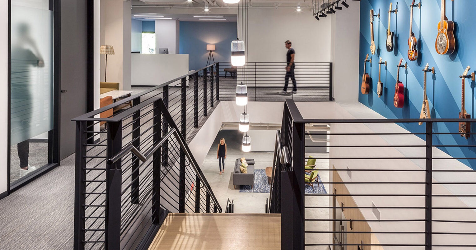Modern office interior featuring a sleek black staircase with metal balusters, connecting two levels. White pendant lights illuminate the space, while a wall-mounted guitar collection adds a creative touch. Open floorplan with glass-partitioned rooms contributes to a spacious and collaborative environment.