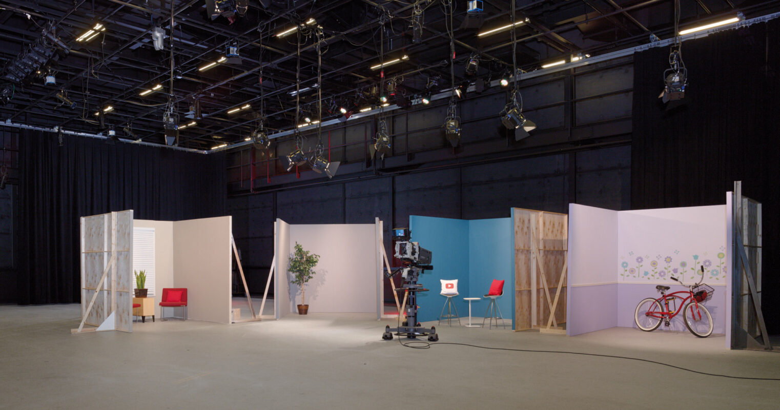 A spacious broadcast studio with high ceilings, equipped with professional lighting rigs and a variety of set pieces including walls, furniture, and props for versatile scene setups.