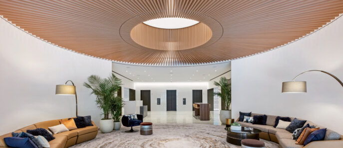 Modern lobby with a concentric patterned marble floor leading to tan sofas accented with blue and gray pillows. Curved wooden slat ceiling with central lighting feature, flanked by tall potted plants and contemporary floor lamps, conveys a warm, inviting atmosphere.