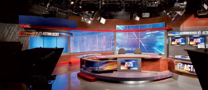 Modern broadcast news studio interior featuring sleek, illuminated desks with geometric design, vibrant blue accent lighting, and multiple flat-screen monitors. Dynamic angular lines contribute to a high-tech, professional ambiance.