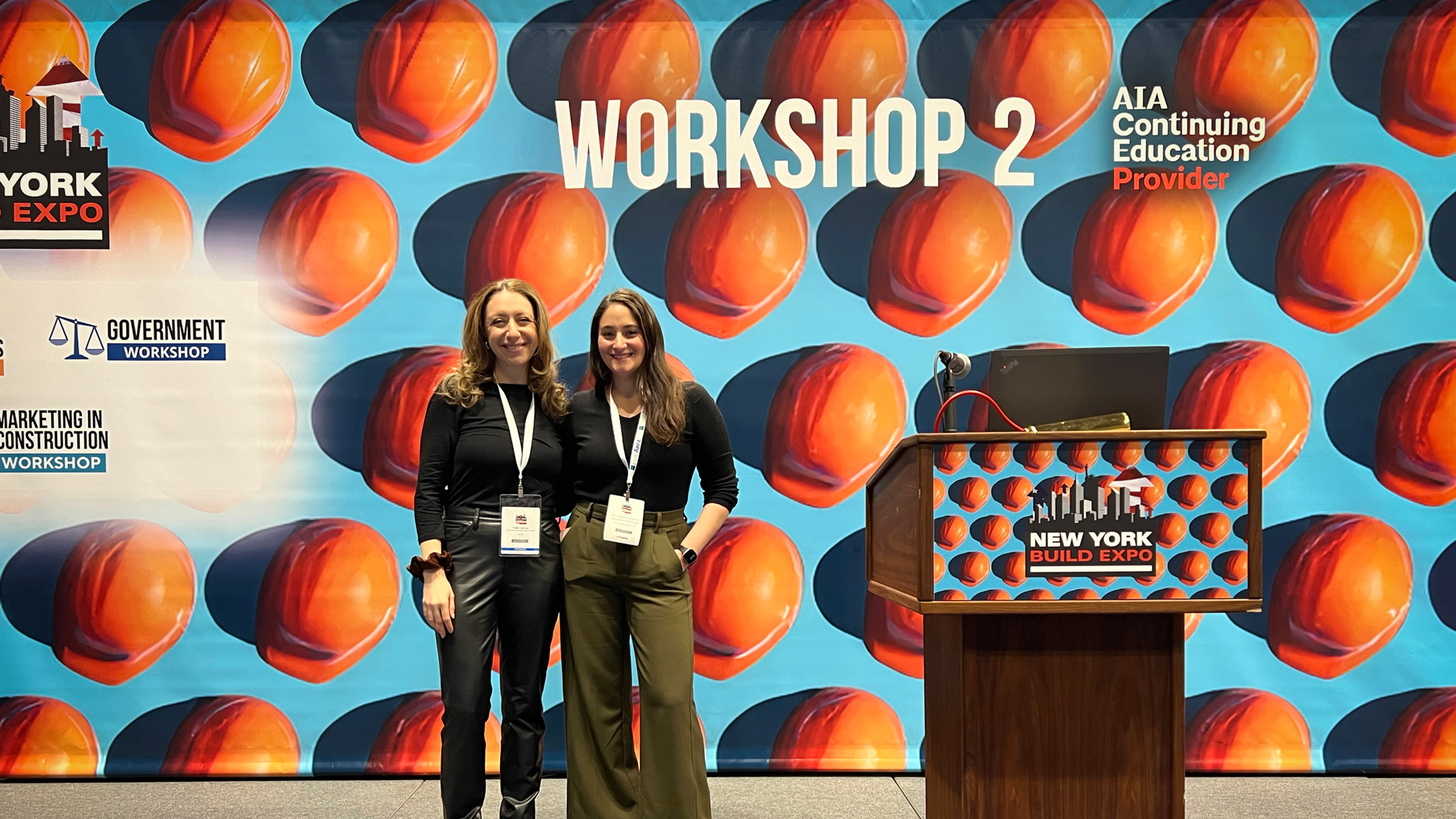 Two smiling women standing in front of a colorful backdrop with large printed hard hats, labeled "WORKSHOP 2" at the New York Build Expo, indicating a focus on construction and architecture.