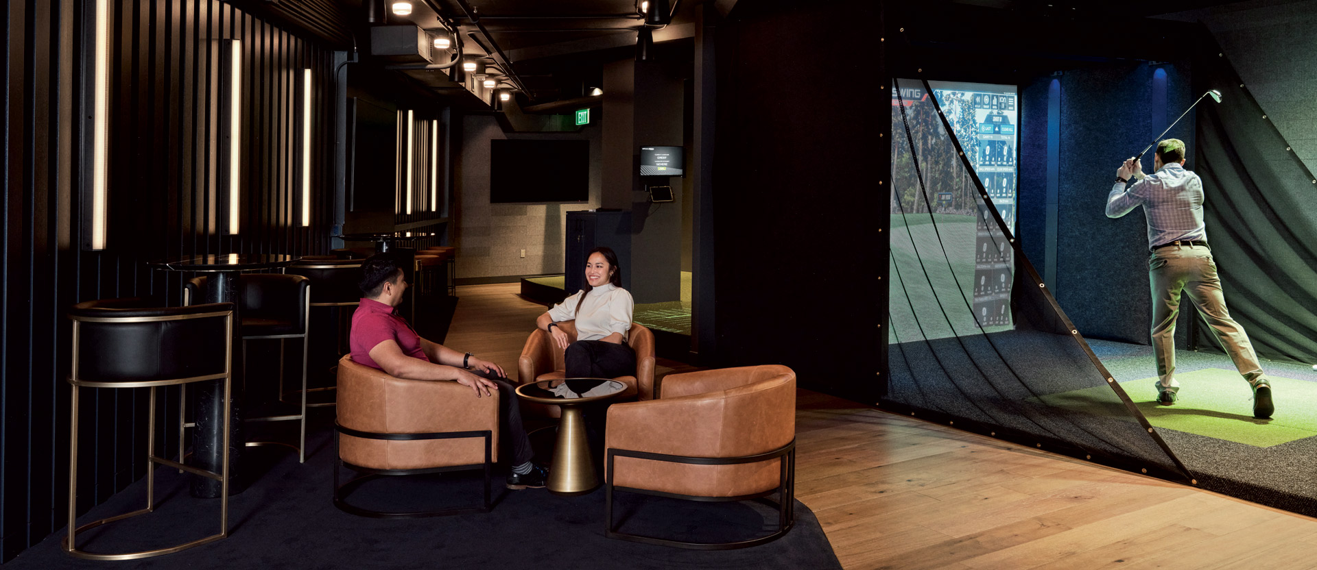 Modern urban-inspired lounge with plush leather seating and industrial-style lighting against a backdrop of dark hues, complemented by a vibrant, full-wall golf simulation screen, offering a dynamic contrast and engaging recreational feature within the space.