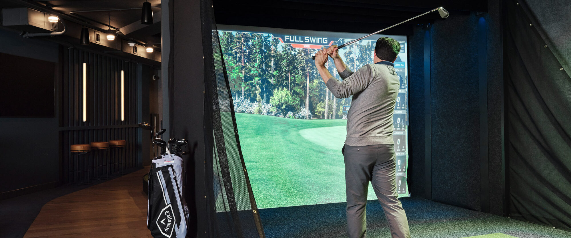 Man practices golf swing in a state-of-the-art indoor simulator space, set against a high-resolution screen displaying a virtual golf course. The room is accented with industrial-style lighting, dark ceiling, and minimalist decor, emphasizing the fusion of technology and comfort in contemporary interior design.