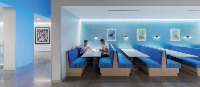 Modern office breakout area featuring vibrant blue upholstered booths, white surfaces, and frosted glass partitions. Strategic lighting accents the space while framed artwork adds a creative touch.