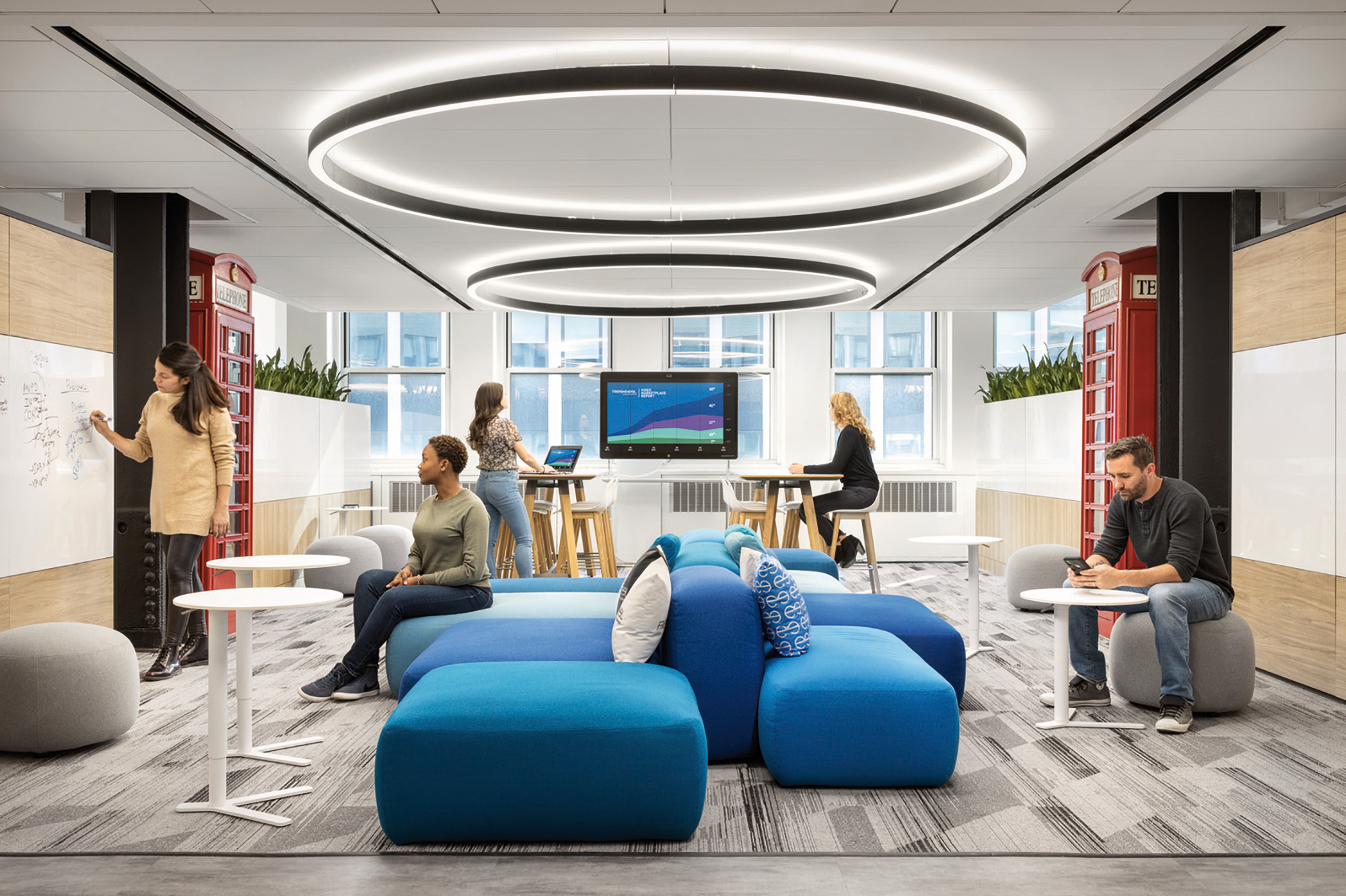 Modern office space featuring an eclectic mix of seating, including bean bags and high stools. An oval overhead lighting fixture mimics the shape of the central seating arrangement, while whiteboards and multimedia screens facilitate collaboration. The design integrates both privacy and openness, with ample natural light.