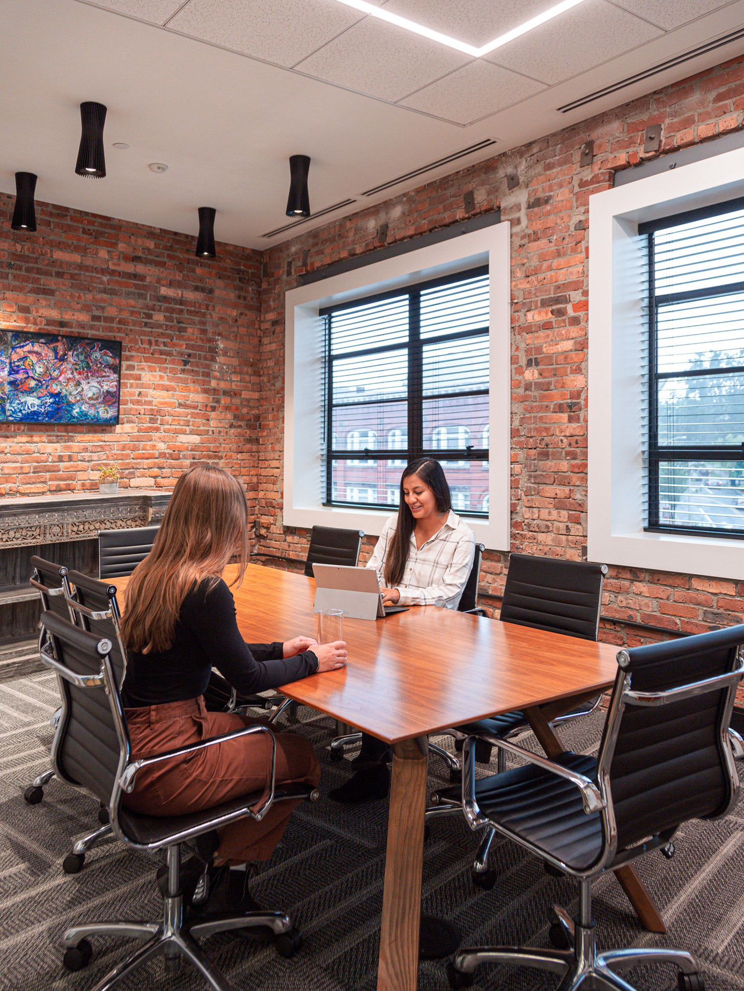 Modern office meeting space with exposed brick wall, contemporary artwork, and large windows allowing natural light. Two professionals are seated at a polished wood table on ergonomic chairs, engaging with a laptop.