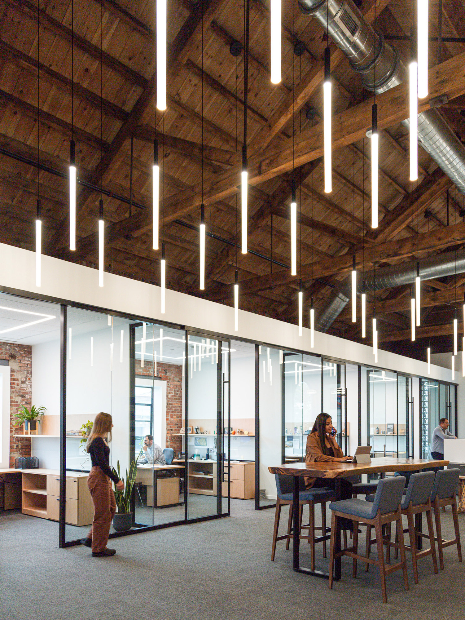 Modern open-plan office space with exposed wooden beams and ductwork, featuring sleek, suspended LED lighting. Glass-walled private offices flank the communal area with employees engaged at workstations and in casual seating.