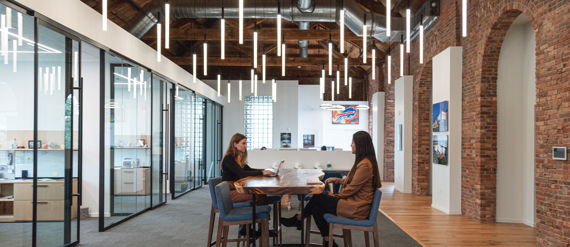 Two professionals sit at a minimalist wooden table in a spacious office with exposed brick walls, wooden beams, and sleek, elongated pendant lights contributing to a modern industrial aesthetic.