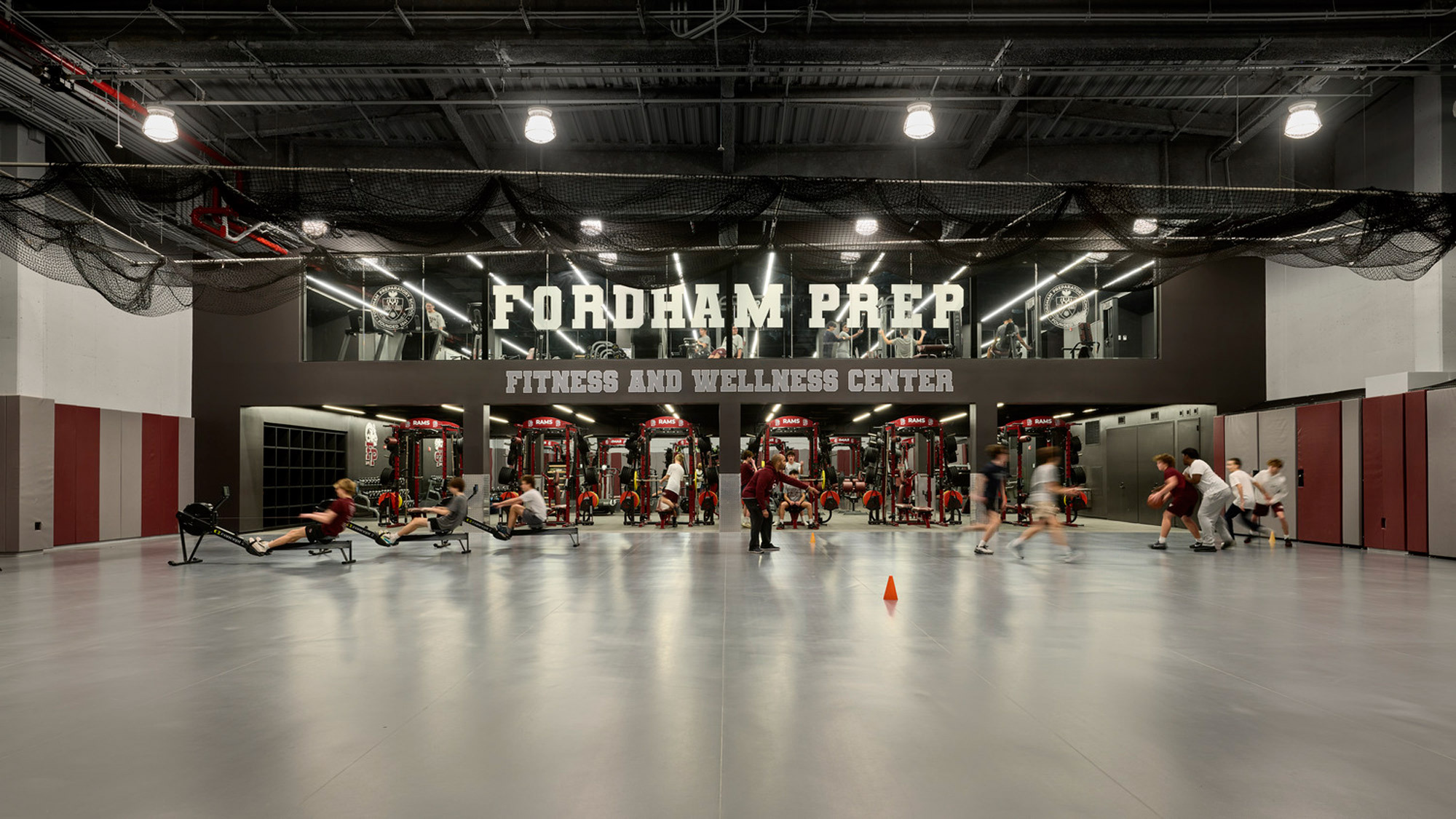 Modern fitness center with high ceilings, exposed ductwork, and industrial lighting. The space has a branded wall with "Fordham Prep Fitness and Wellness Center" and is equipped with cardio and weightlifting machines, balancing the aesthetics with functionality.
