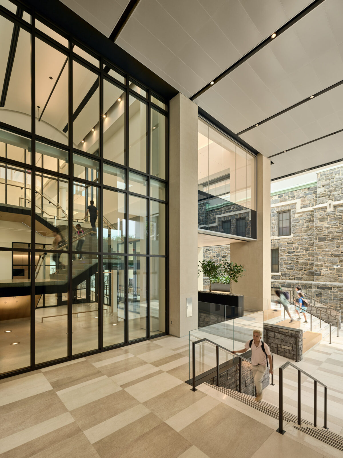 Modern interior featuring a double-height atrium with a checkered floor pattern. Glass partitions provide transparency, complementing the clean lines of the steel staircases. Stone walls add texture, enhancing the space's blend of industrial and natural design elements.