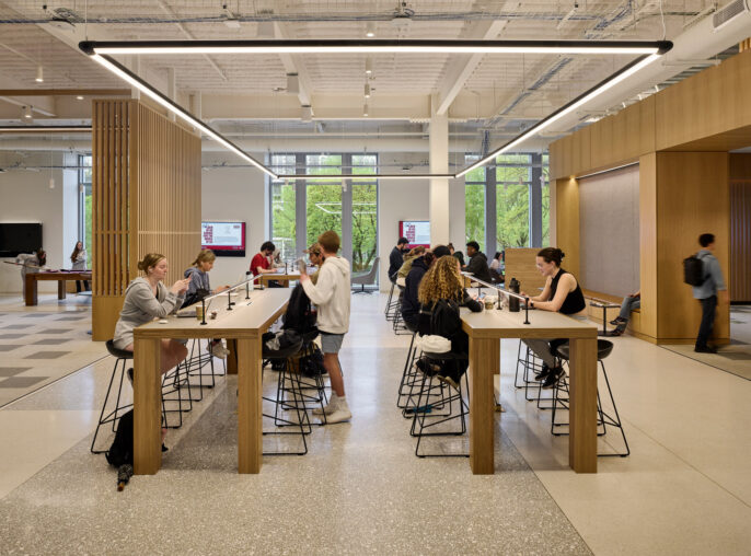 Modern open-plan workspace featuring a central communal table with overhead linear pendant lighting. Individuals work at spaced intervals, surrounded by wood-paneled walls and exposed ceiling infrastructure, which provides an industrial yet refined aesthetic.