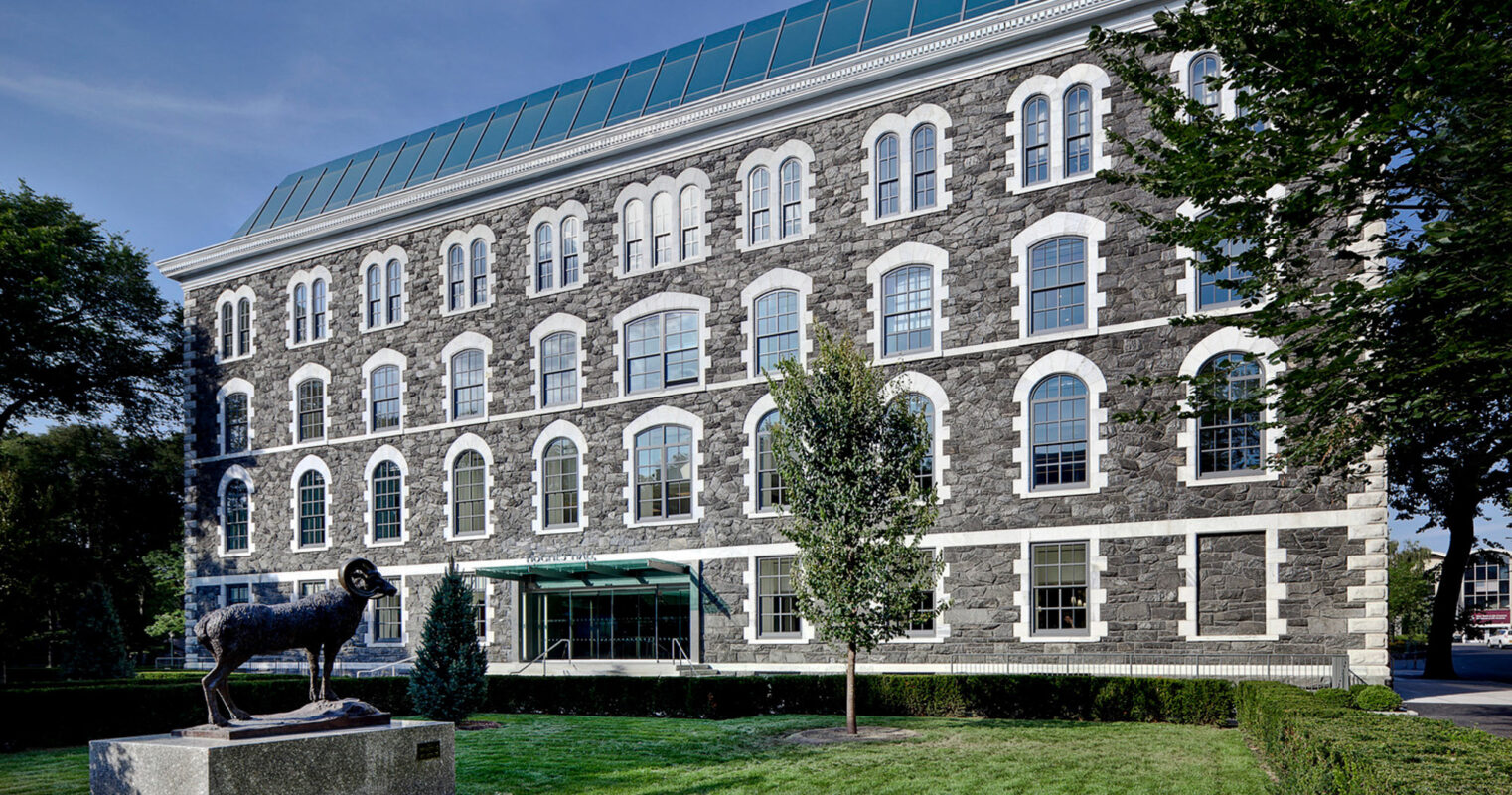 Exterior of a renovated historical building, showcasing a well-preserved stone facade, symmetrically aligned arched windows, and a modern glass rooftop addition that contrasts with the traditional architecture below. A sculptural art piece adorns the manicured front lawn.