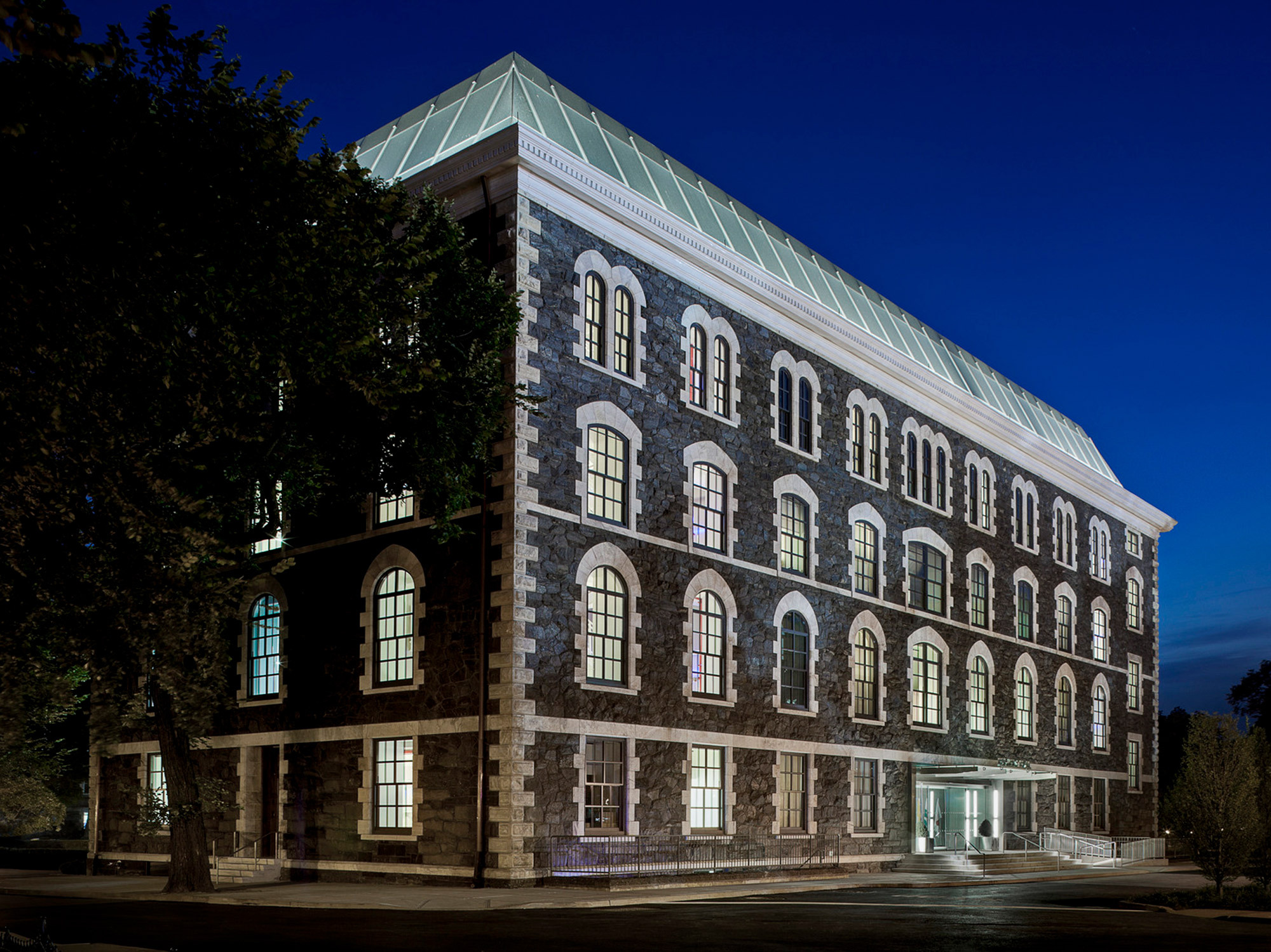 Twilight highlights the stone facade and pronounced arches of a renovated historical building, featuring a modern glass roof addition that blends old architectural charm with contemporary design.