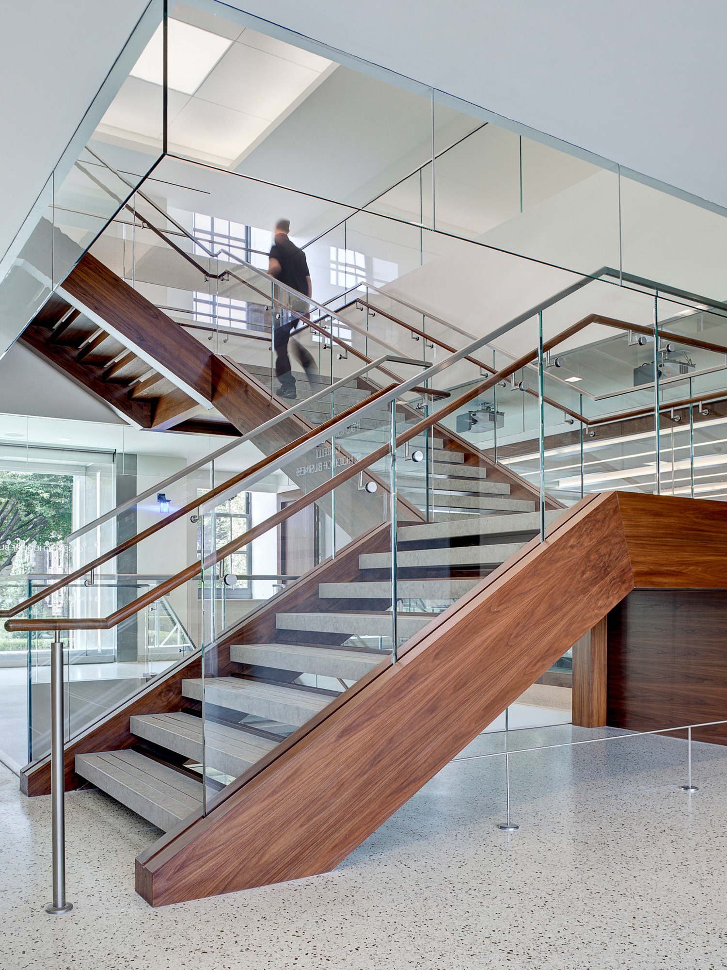 Open-concept staircase with floating wooden steps flanked by glass balustrades, enhancing spatial connectivity. The structure features brushed steel handrails and a warm, rich walnut finish, contributing to a modern, airy ambiance within a professional or residential setting.
