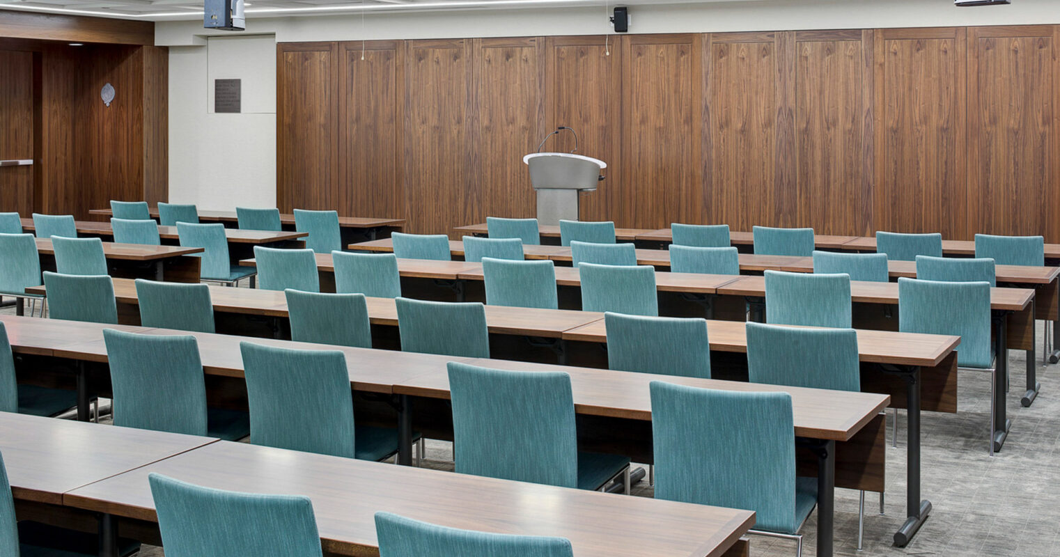 Modern conference room featuring rows of turquoise chairs, dark wood-paneled walls, and a sleek lecturer's podium. Overhead, linear lighting complements the room's clean lines.
